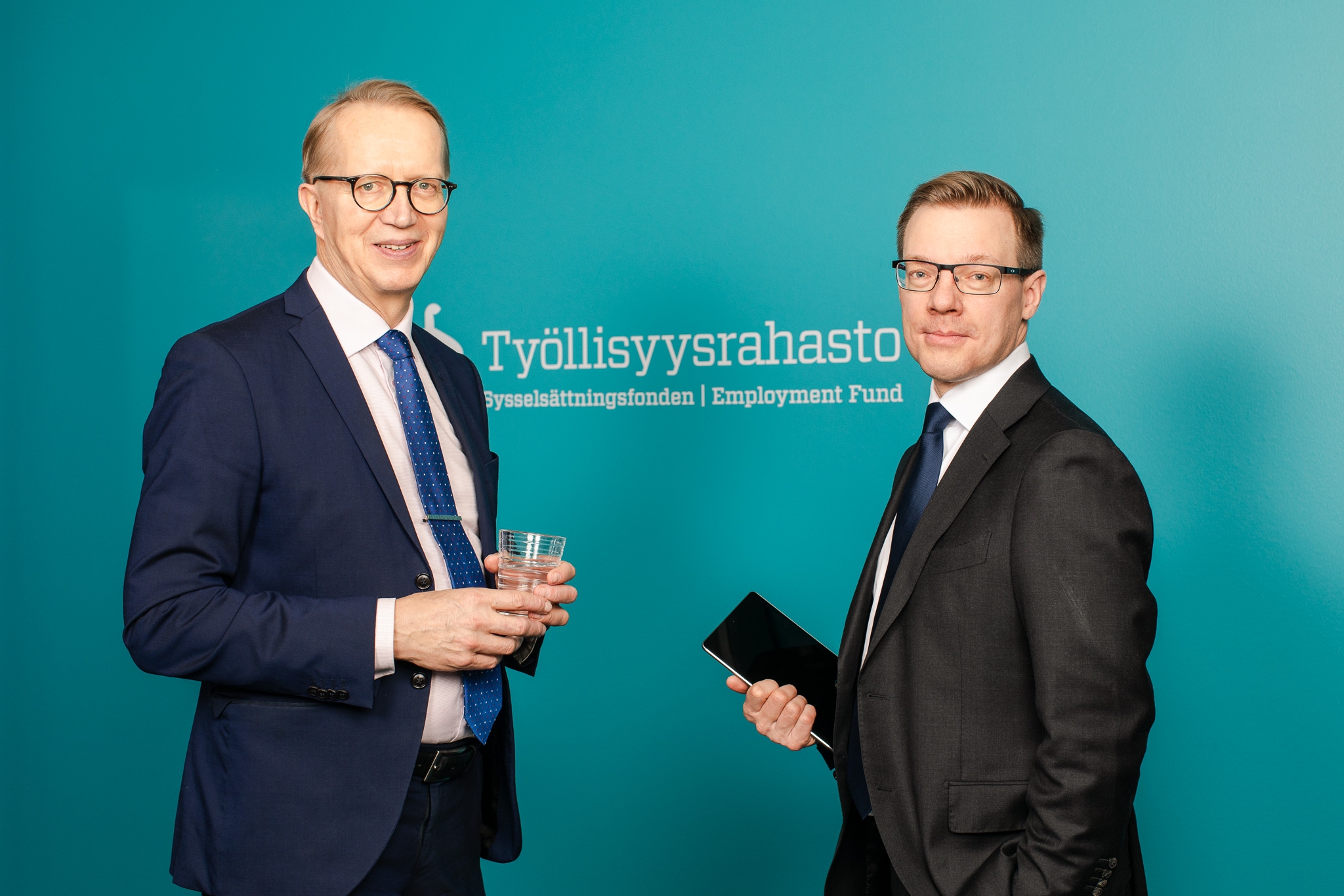 Janne Metsämäki och Karo Nukarinen, står mot varandra i kostymer och tittar mot kameran med ett leende.